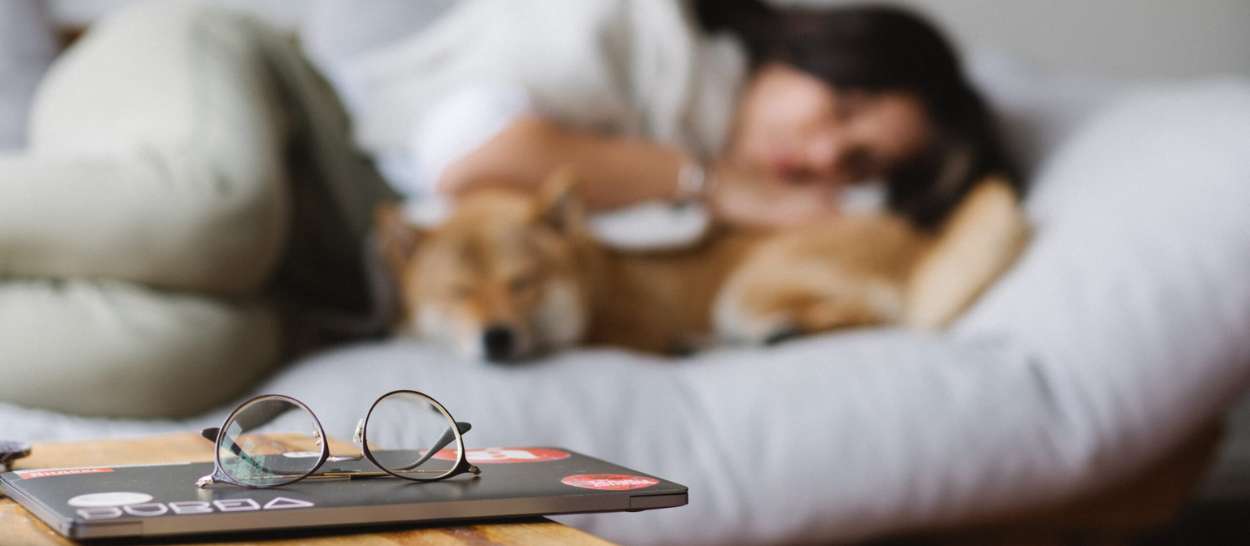 A woman resting on the bed with her dog and her glasses resting on the table